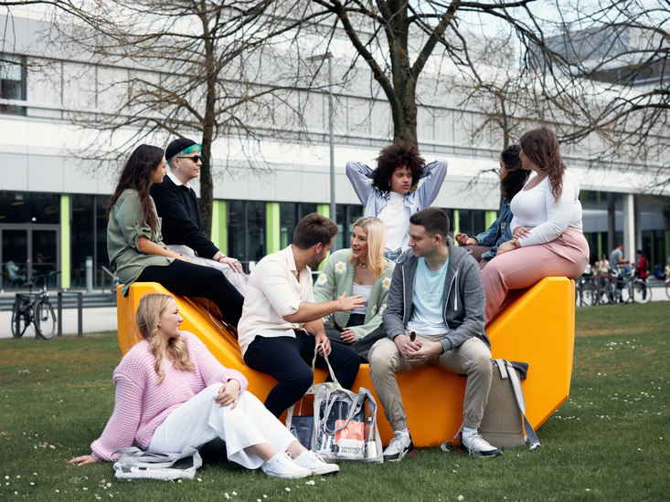 Eine Gruppe Studierender sitzt auf einer orangefarbenen Sitzgelegenheit vor dem X-Gebude.