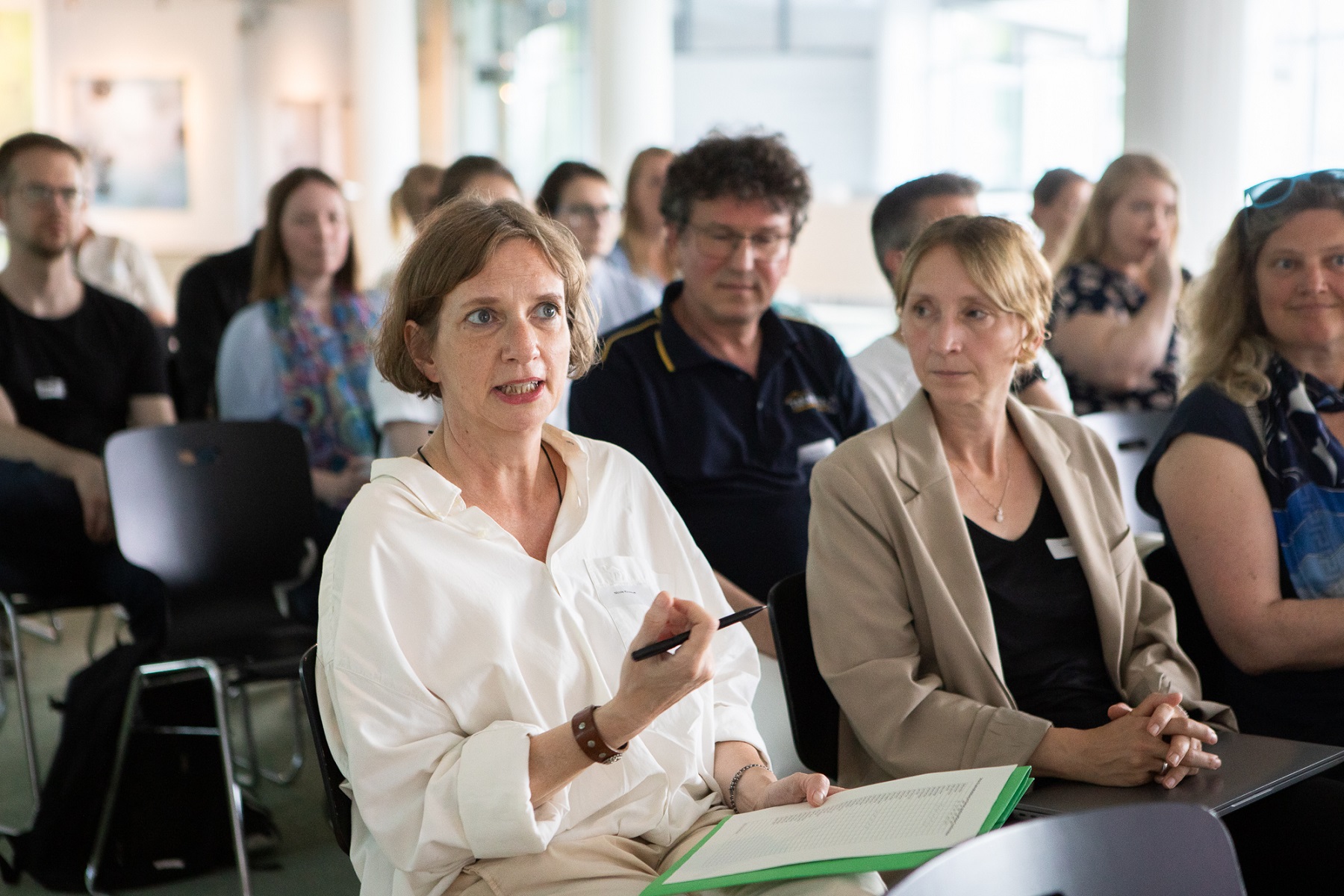 Es sind Personen zu sehen, die in Reihen hintereinander sitzen. Vorne links ist eine Frau zu erkennen, die gerade konzentriert spicht, die anderen Personen gucken zu ihr. 