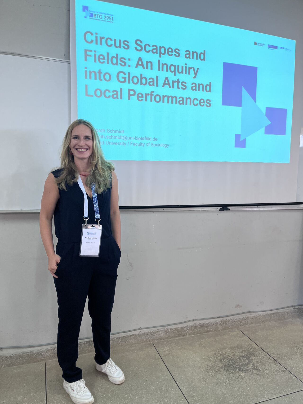 Elisabeth Schmidt in front of projector screen showing the title page of her presentation: "Circus Scapes and Fields: An Inquiry into Global Arts and Local Performances"