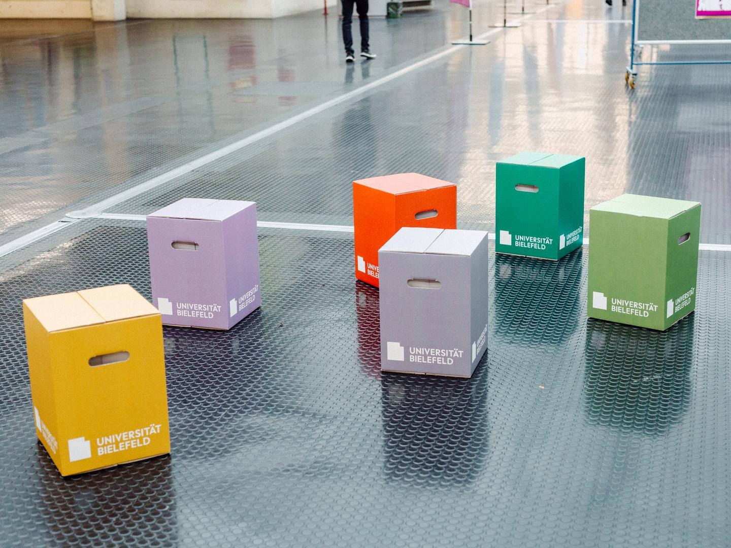 Eine Gruppe bunter Hocker aus Pappe in einer Halle. 