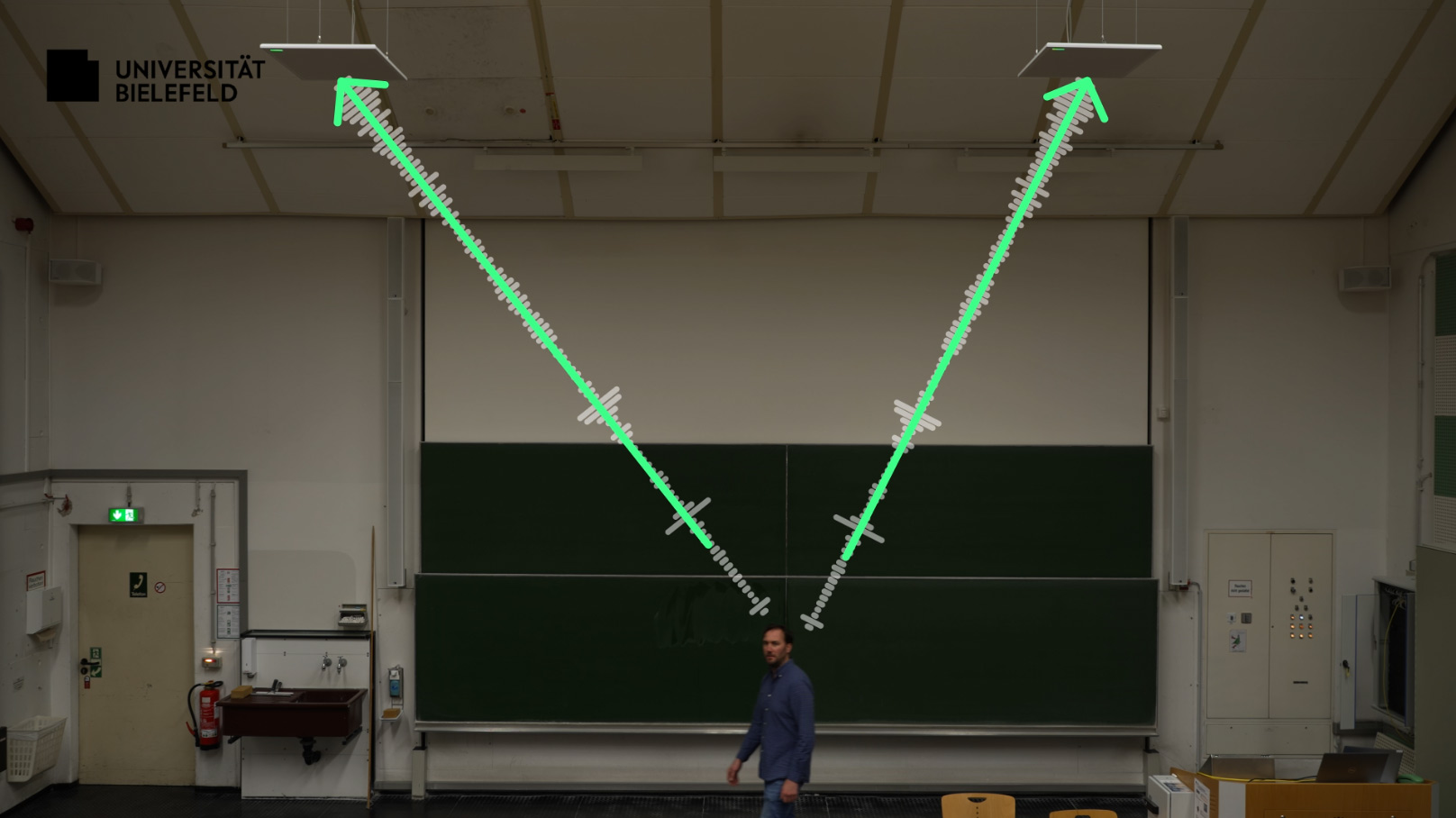 Person steht vor der Tafel im Hörsaal. Grafisches Pfeile zeigen auf die Deckenmikrofone.