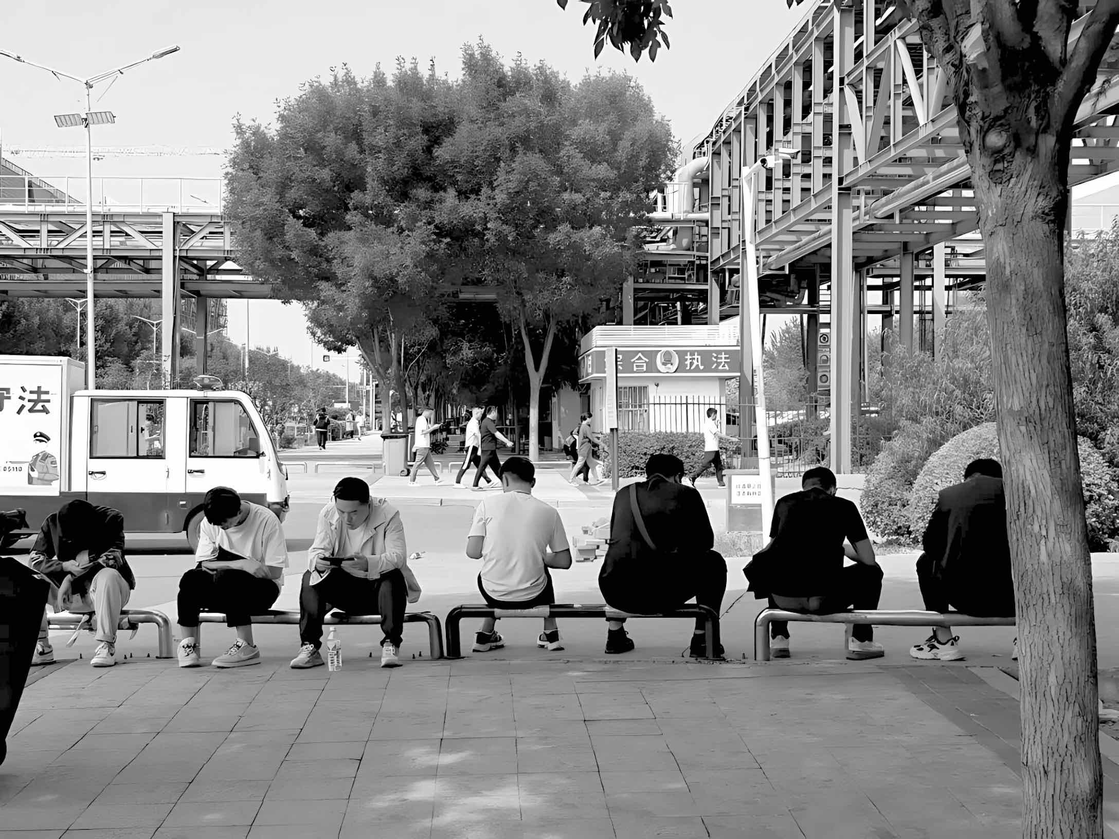A row of young people sitting on benches outdoors, most looking down at their phones, with industrial structures and trees in the background