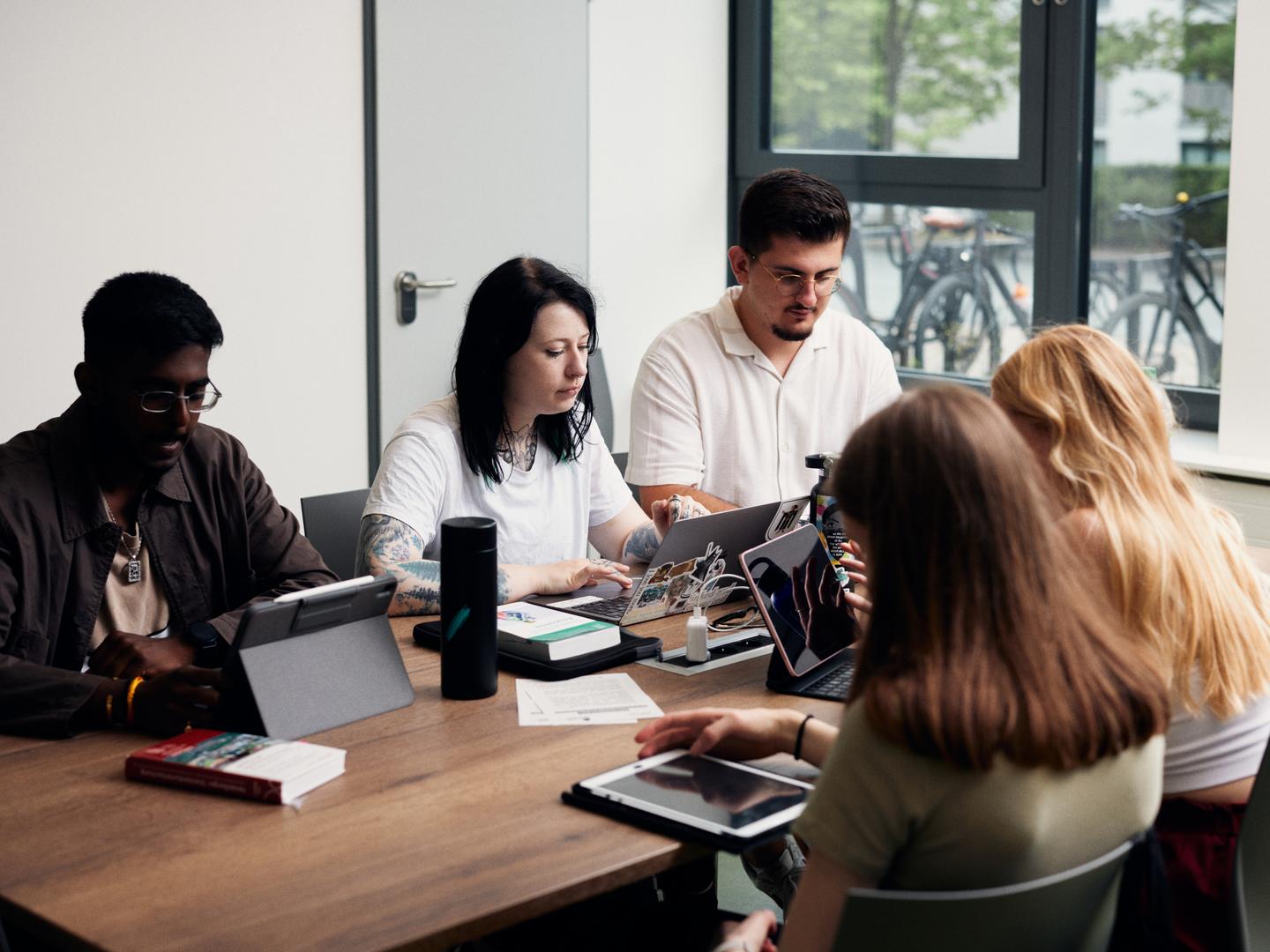 Auf dem Bild ist eine Gruppe von Studierenden zusehen, die im Gemeinschaftsraum an einem Tisch sitzen und am Laptop arbeiten bzw. lernen.