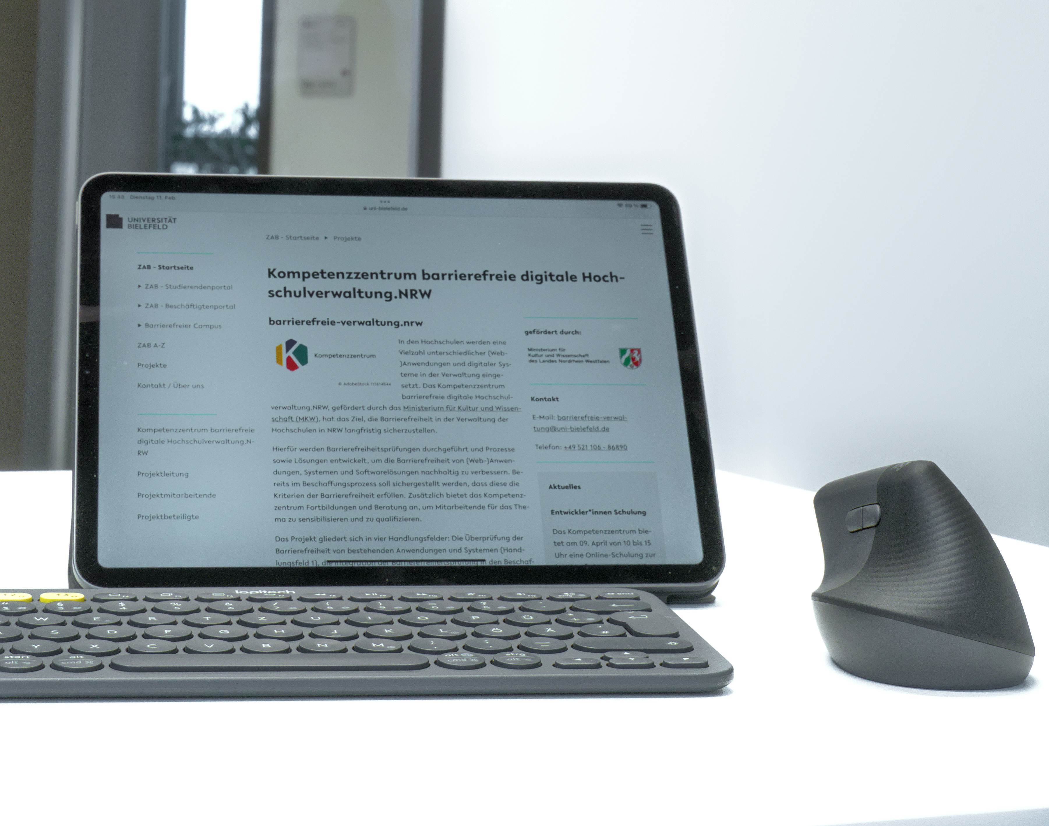 Foto: auf einem Schreibtisch ist ein Tablet, aus dem die Webseite des Kompetenzzentrums geöffnet ist, eine Tastatur und eine Computermaus. Im Hintergrund ist eine geöffnete Tür.