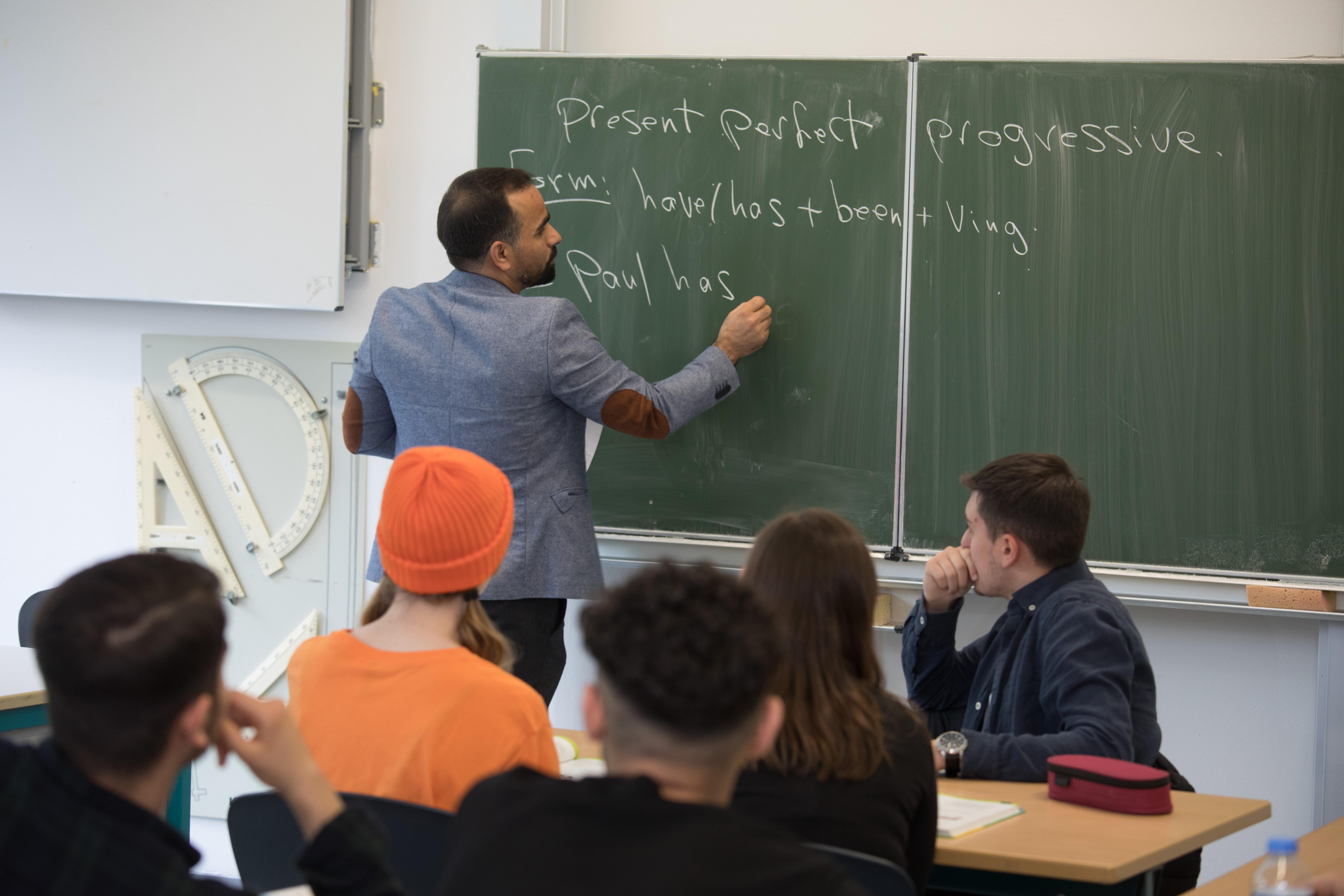 Lehrer schreibt den englischen Beispielsatz "Paus has..." mit Kreide an die Tafel. Im Vordergrund sitzen 5 Schlerinnen und Schler, deren Blicke auf die Tafel und Lehrer gerichtet sind.