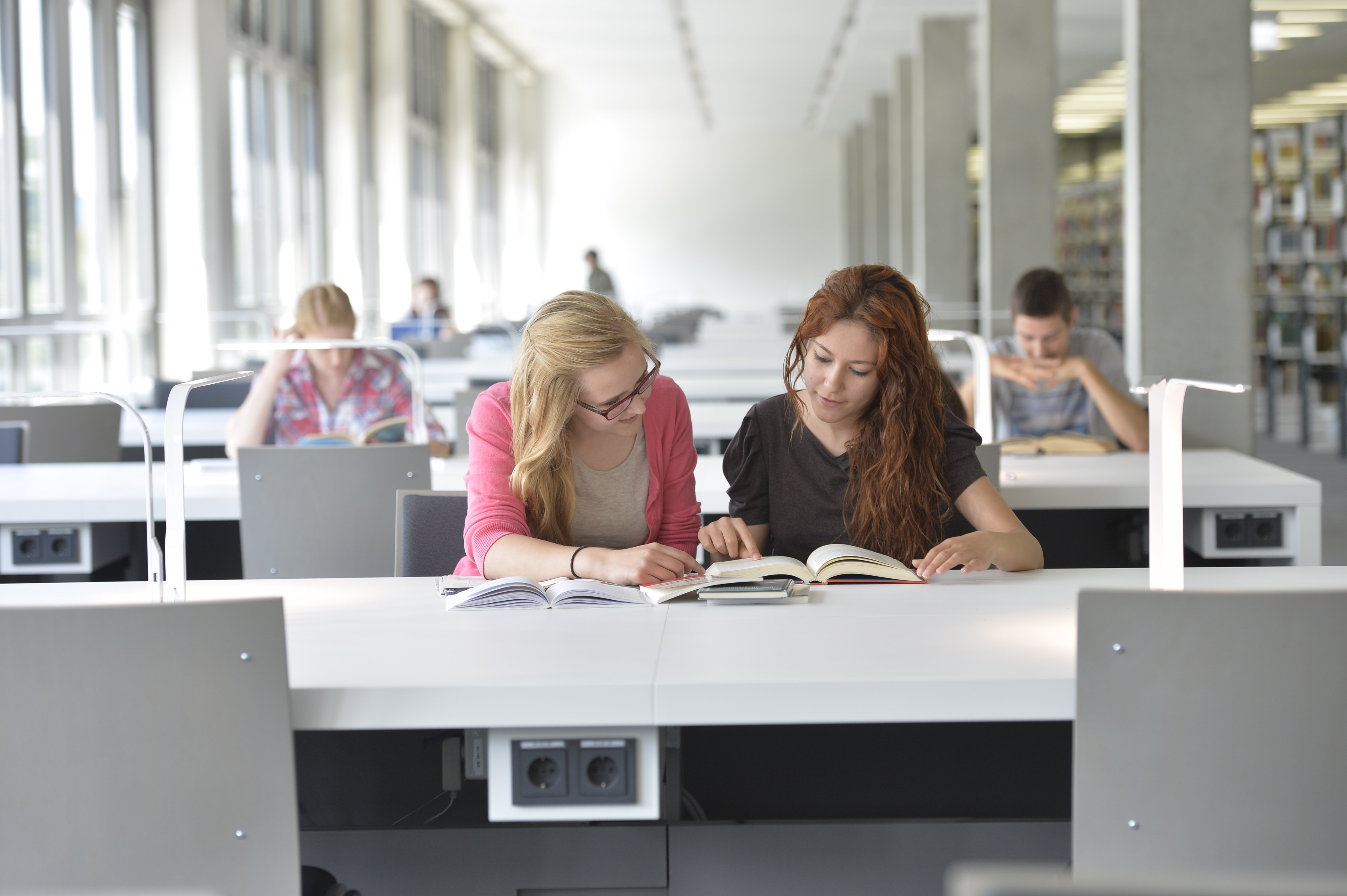 Zwei Studentinnen sitzen gemeinsam über Bücher gebeugt an einem Tisch in der Bibliothek