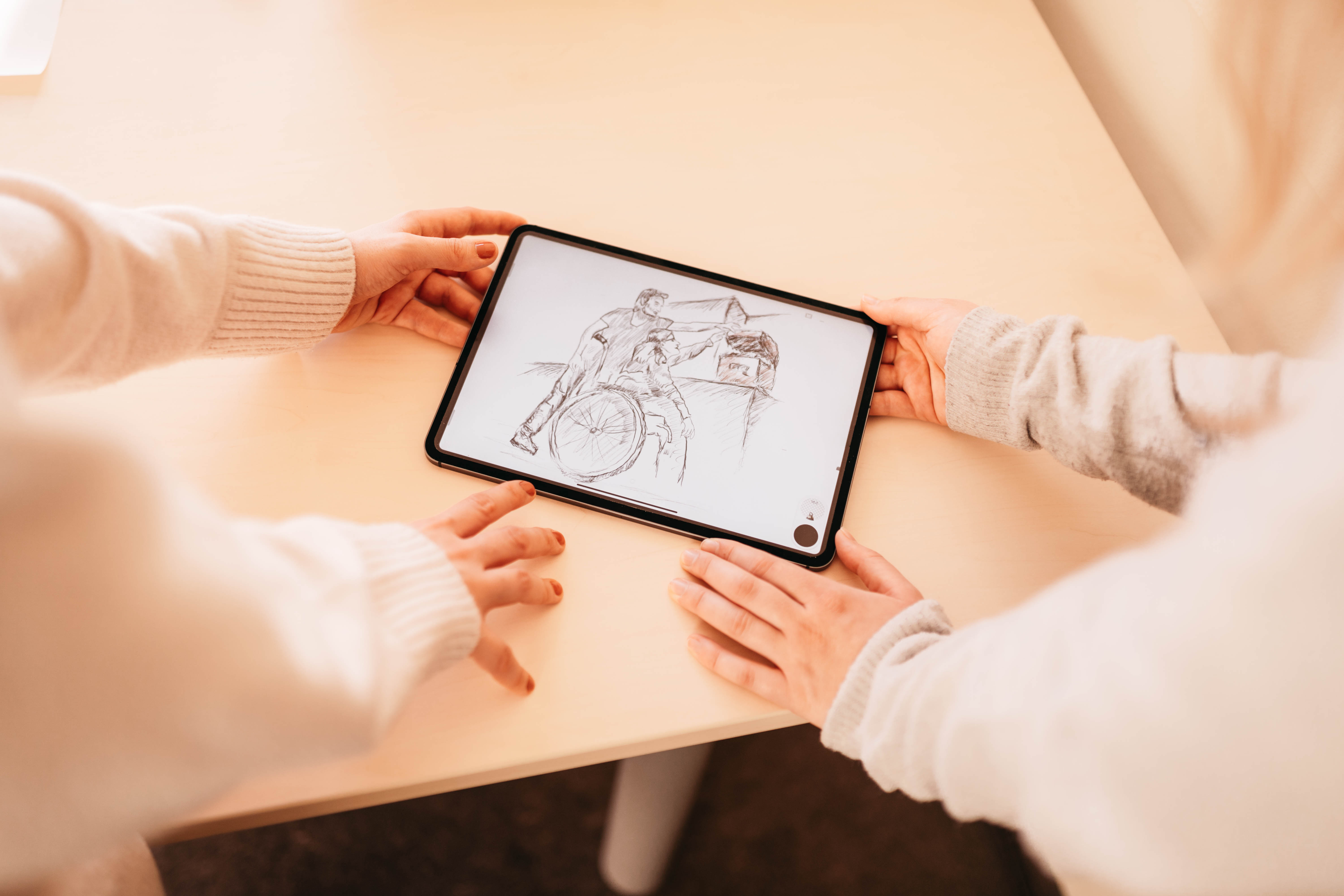 Two staff, people are holding a tablet on which a drawing with a wheelchair user can be seen