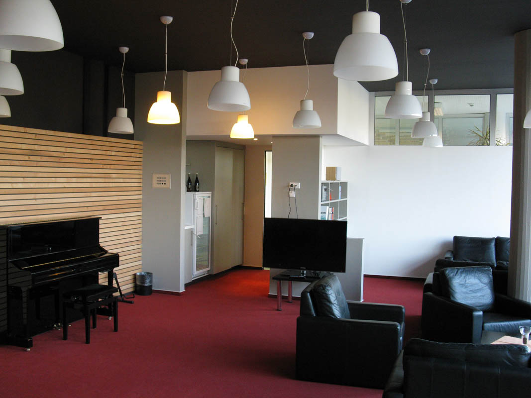 The fellow room with its wine red carpet, black sofas, a piano, a wooden wall and lots of ceiling lamps