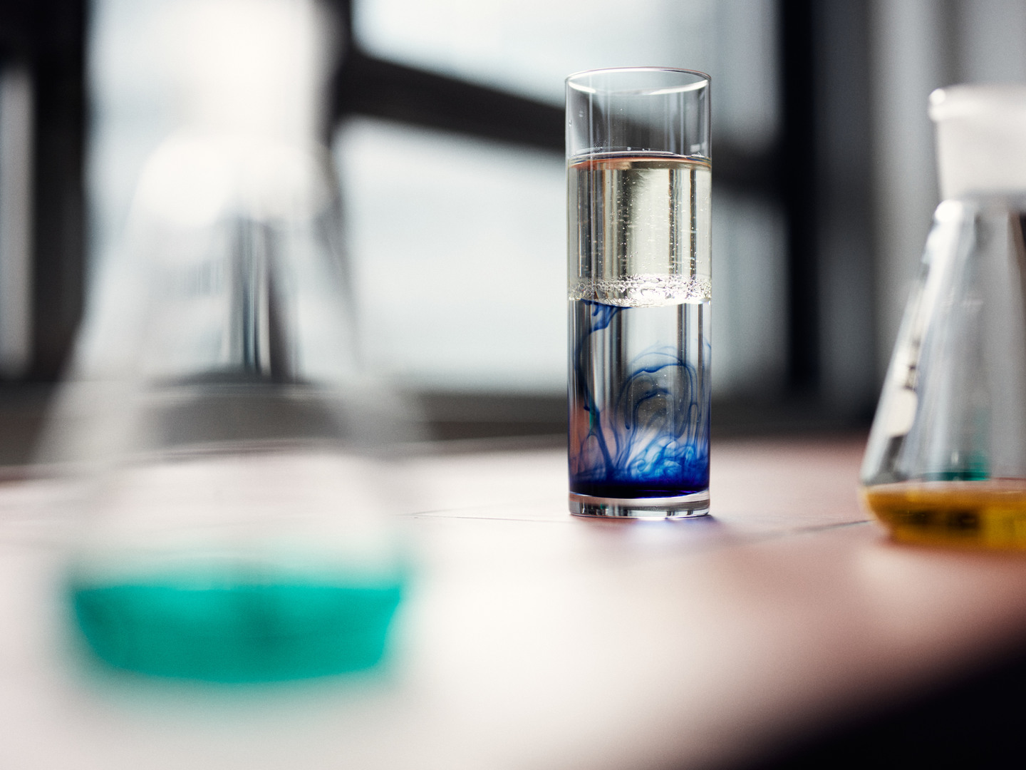 Ein Glas in dem Öl auf Wasser schwimmt mit blauer Farbe unten im Wasser. Rechts und links sind unscharfe Erlenmeyerkolben.