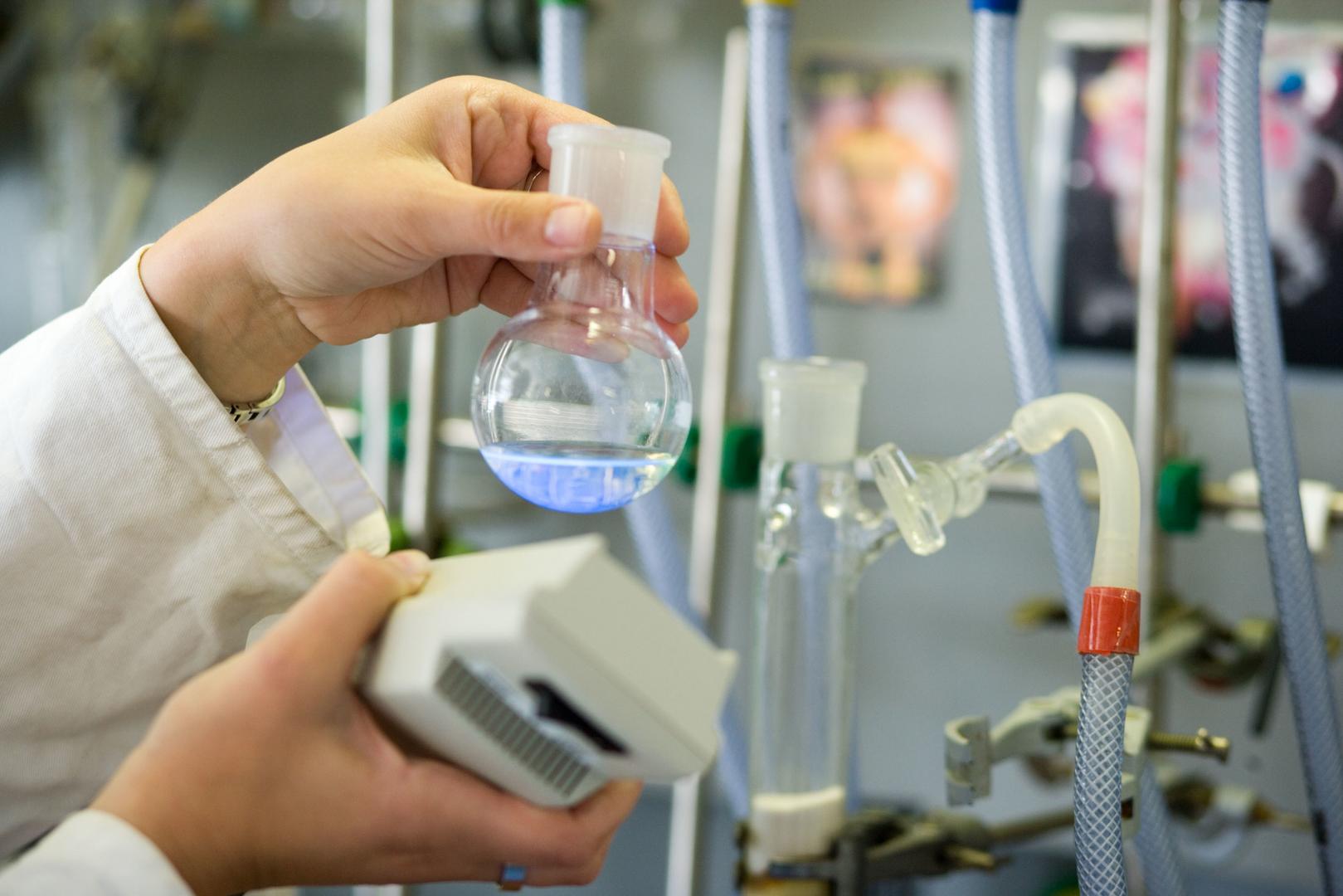Eine Person im Labor hält vor einer Versuchsapparatur einen Kolben mit blauer leuchtender Flüssigkeit in der Hand.