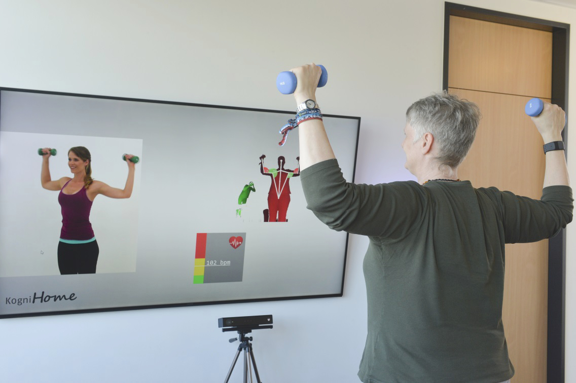 A standing person with dumbbells imitates the movements of an avatar on a screen