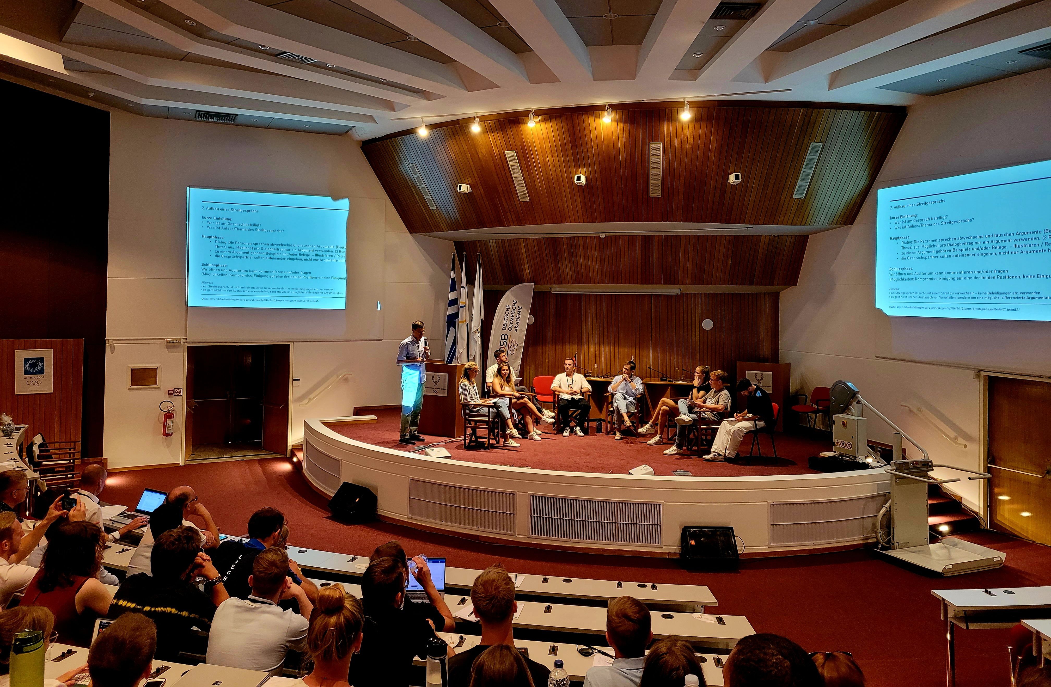 Acht Personen sitzen auf einem Podium und eine Person spricht in ein Mikrofon vor der Projektion einer Präsentationsfolie