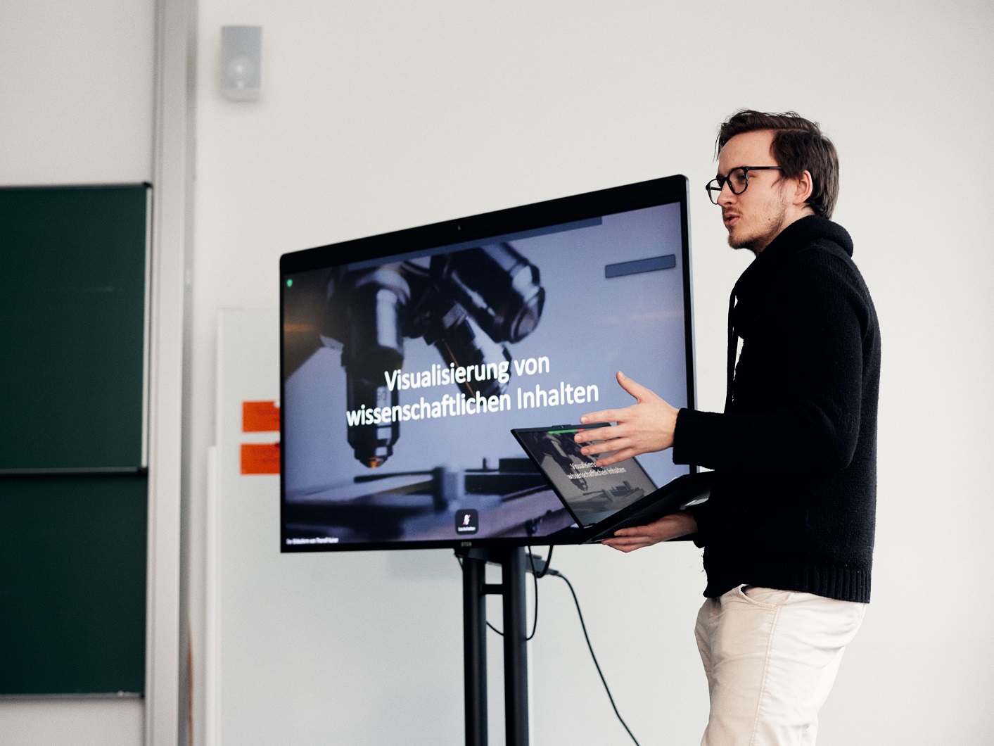 Ein Mann hält einen Laptop und stellt eine PowerPoint-Präsentation vor, die neben ihm auf einem großen Bildschirm angezeigt wird