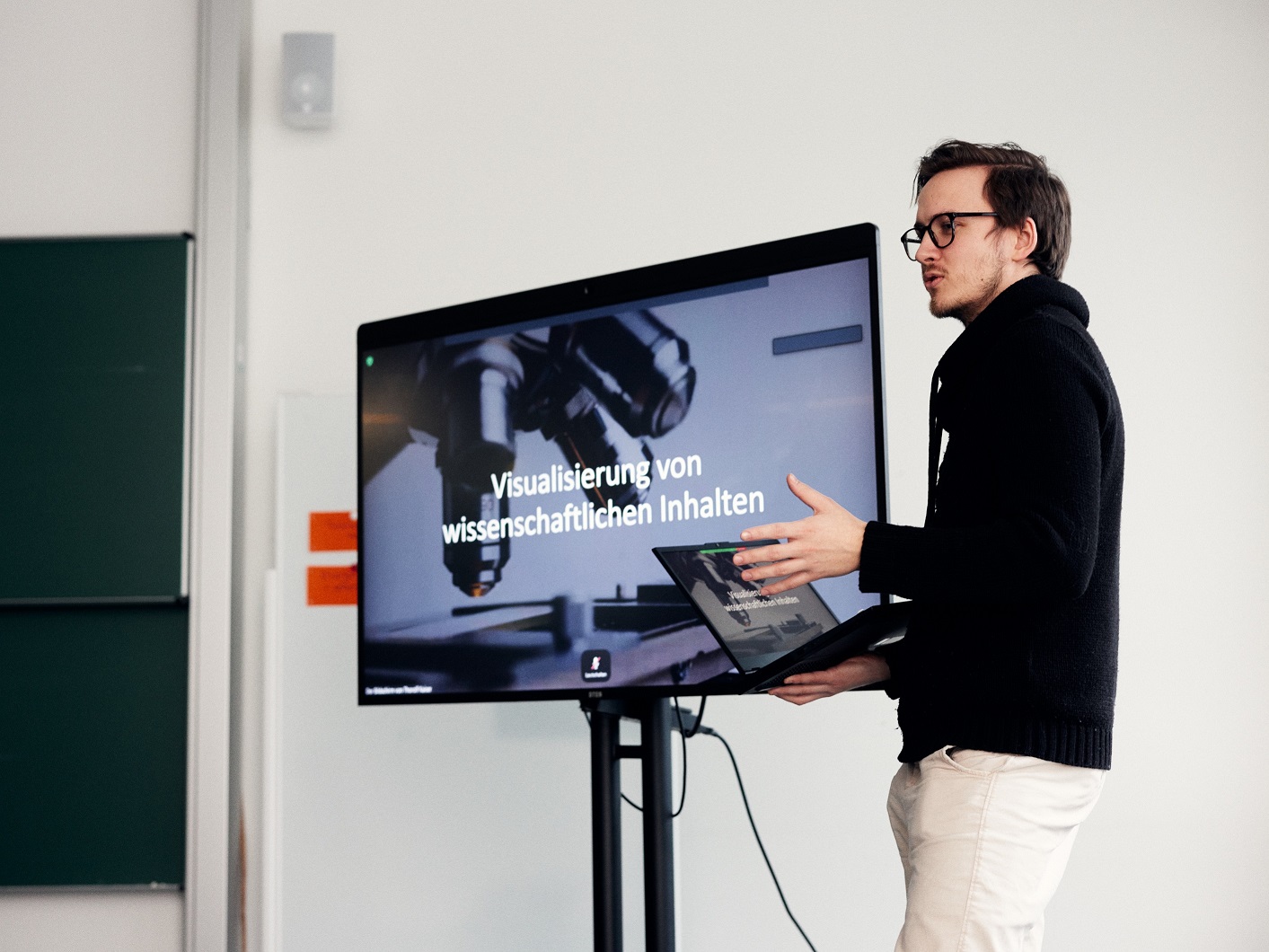 A man holds a laptop and presents a PowerPoint presentation that is displayed next to him on a large screen