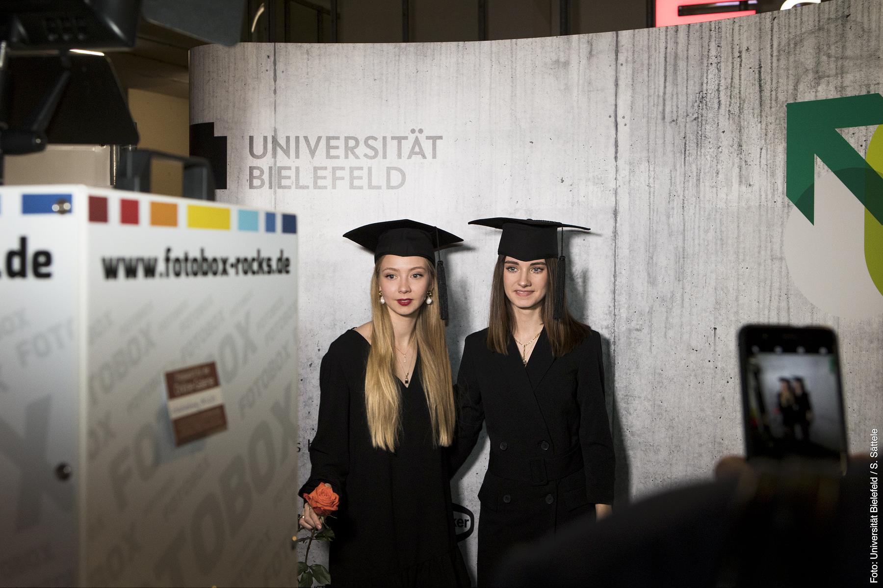 Zwei Absolventinnen vor einem Fotohintergrund im Uni-Design
