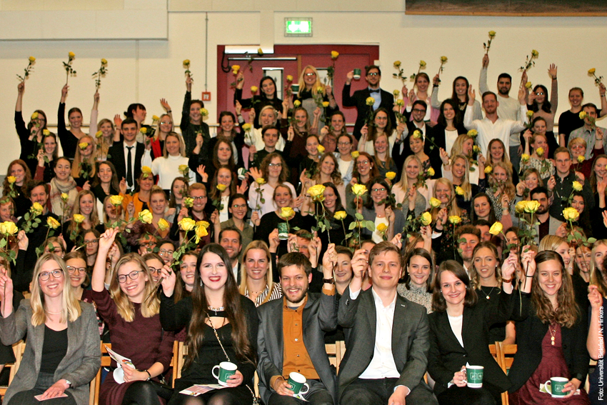 Gruppenfoto der Absolvent*innen, die alle eine gelbe Rose in die Luft halten