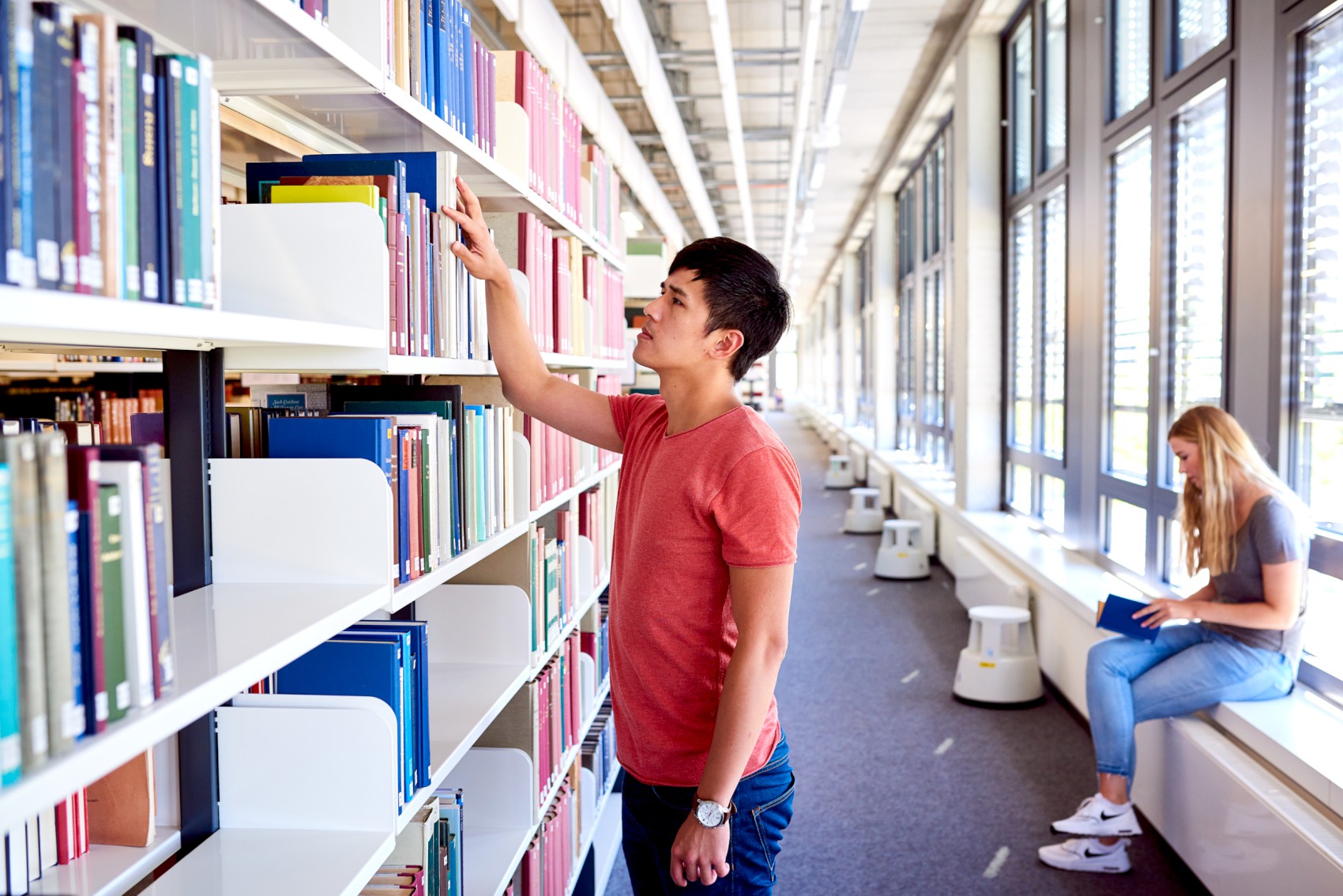 Students in the library, student at a shelf, a female student sitting and leafing through a book