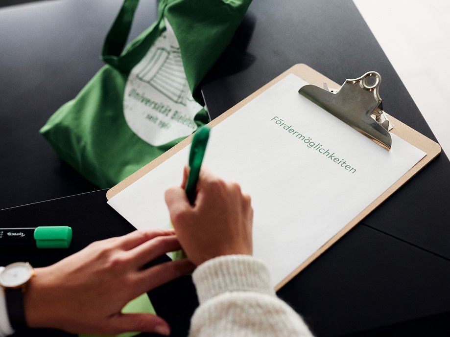 Eine Person schreibt auf ein Blatt auf einem Klemmbrett, auf dem "Fördermöglichkeiten" gedruckt ist. Neben dem Klemmbrett liegt ein grüner Jutebeutel mit dem Universitätslogo.