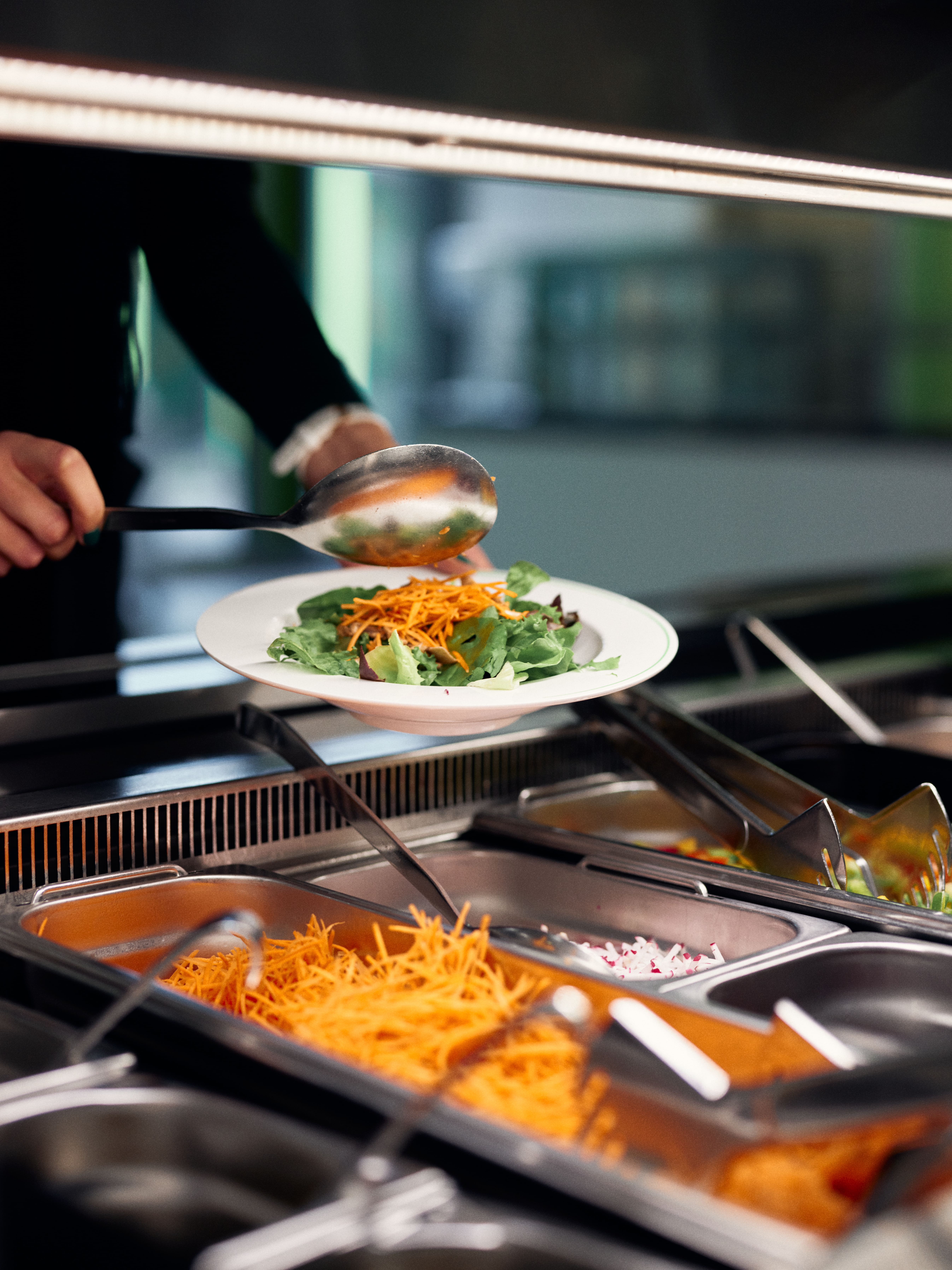 Person befüllt sich einen Teller mit Salat an der Salatbar in der Mensa. 