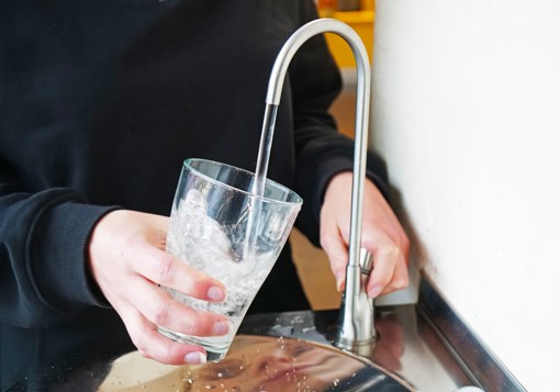 Ein Glas wird von einer Hand gehalten und mit Wasser an einem Trinkwasserspender befüllt.
