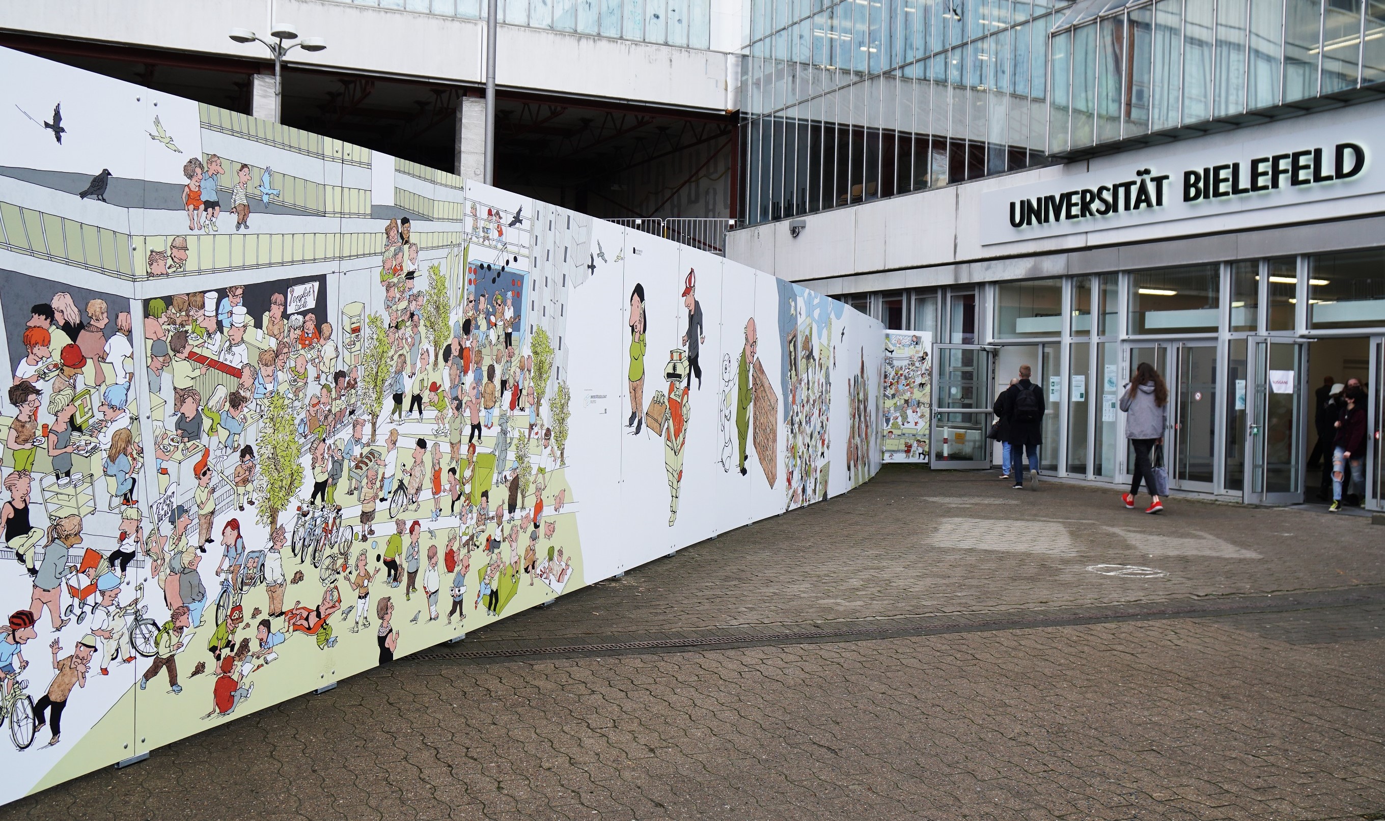 Figuren aus dem Jubiläumsbuch "Unigewimmel - 50 Jahre Universität Bielefeld" von Peter Menne schmücken den neuen Bauzaun auf dem Interimsvorplatz des Universitätshauptgebäudes