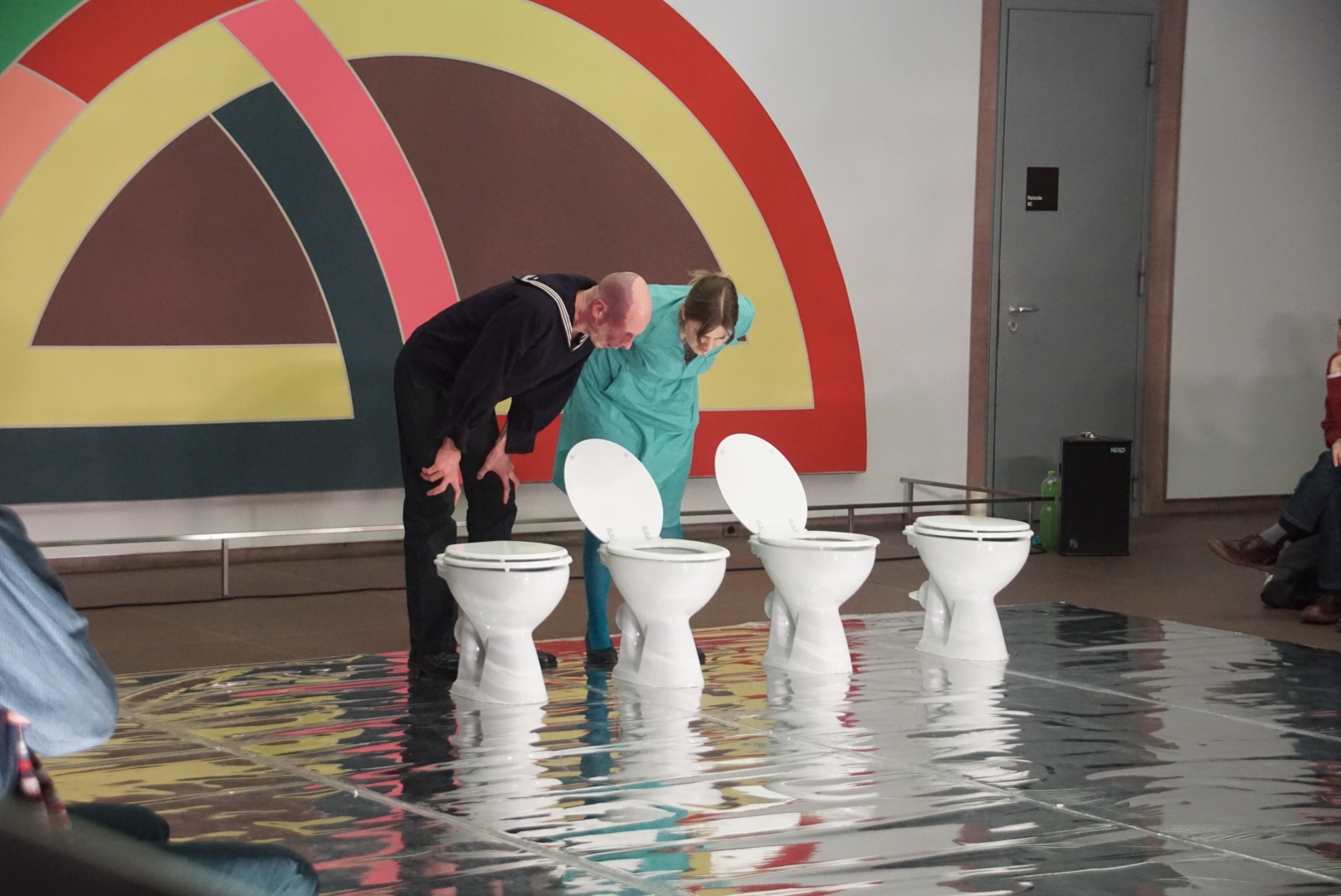 Zwei Künstler*innen beugen sich in einer gespielten Szene über Toiletten, die frei im Raum stehen