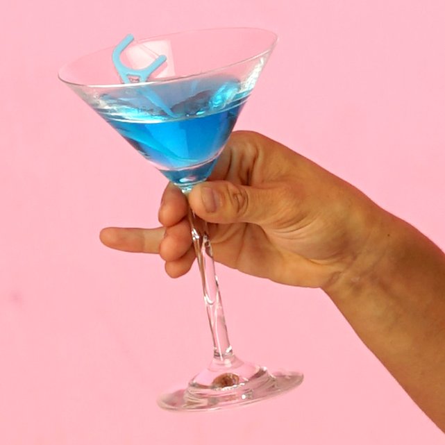 Eine Hand hält ein Martiniglas gefüllt mit einer blauen Flüssigkeit. Im Glas schwimmt ine Zahnseidestäbchen.