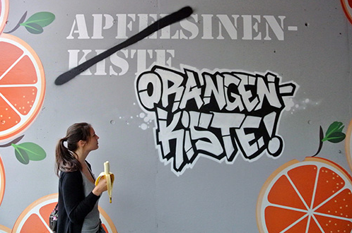 Frau mit einer Banane in der Hand liest die Aufschrift auf einer Wand: Das Wort Apfelsinenkiste ist durchgestrichen und durch Orangenkiste ersetzt.