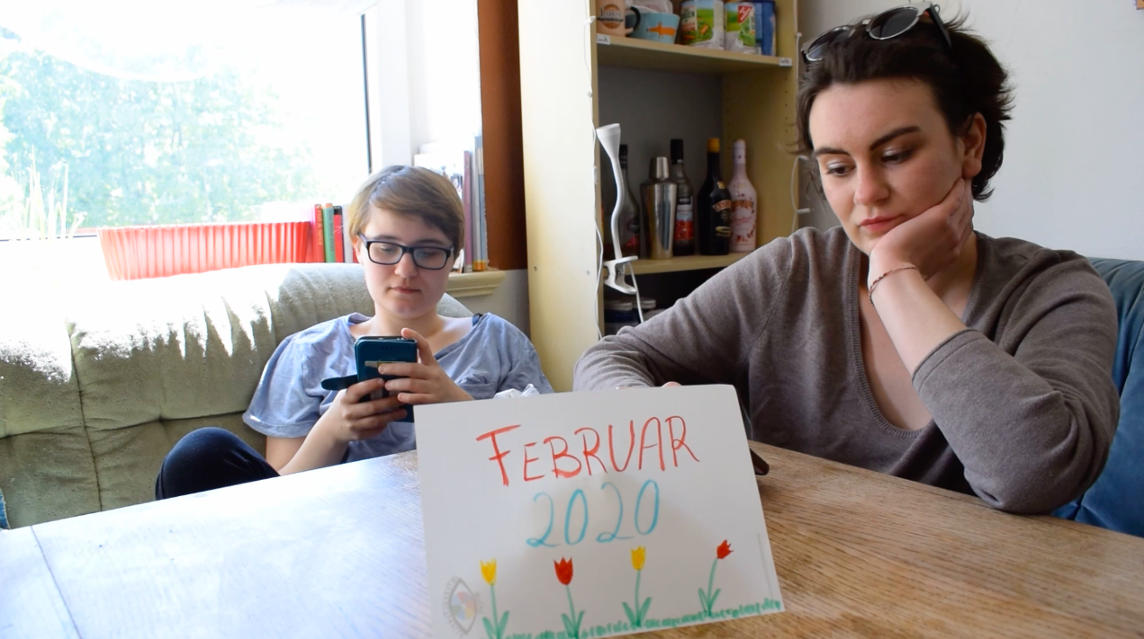 Zwei junge Frauen sitzen an einem Tisch, beide schauen auf ihr Handy. Auf dem Tisch steht auf einem Zettel "Februar 2020"