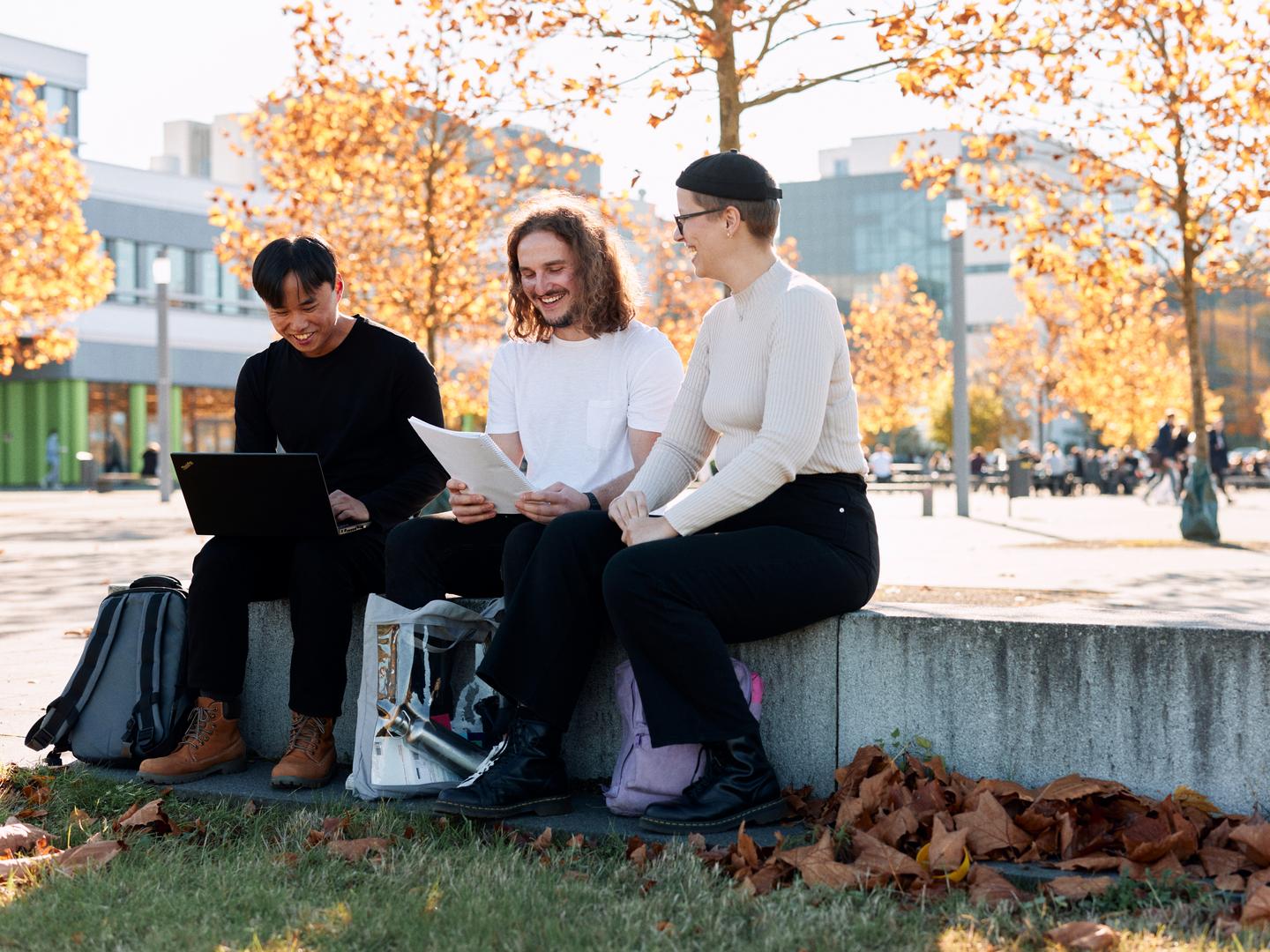 Three students are sitting on a little wall on campus, studying papers and talking. One young man has a laptop on his lap, another is looking at a notepad.