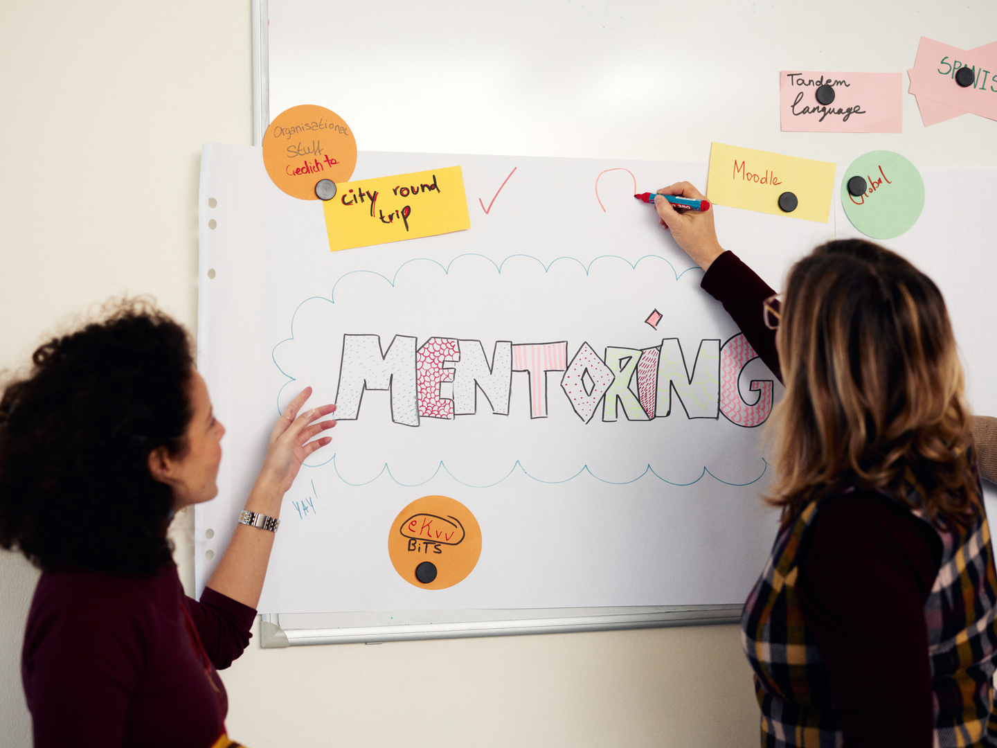 Zwei Doktorandinnen schreiben Mentoring auf ein PLakat an der Wand