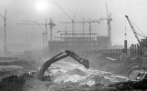 The construction site of the main university building in 1972. In the foreground, an excavator is digging in the earth. In the background, the beginning shell of the main building can be seen with numerous cranes.